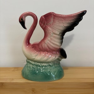 May include: A ceramic flamingo figurine featuring a curved neck, a black beak, and wings extended upward. The body is shaded in shades of pink and white, with black accents on the wing tips. The base is molded to resemble green grass.