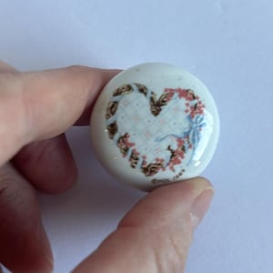 May include: A round white ceramic knob featuring a central heart design composed of a blue checkered pattern, pink floral accents, and brown ribbon details. The text CHOL 1989 is printed in small black letters near the bottom edge of the knob.