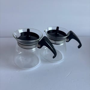 May include: Two clear glass coffee carafes featuring black plastic lids with central knobs and black plastic side handles. Each carafe has a silver-toned metal band encircling the neck, connecting the handle to the glass body.