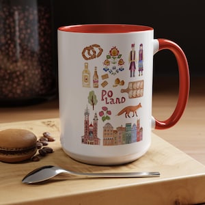 May include: A white ceramic mug with a red interior and handle, featuring colorful illustrations of Polish cultural elements. The design includes food, buildings, and the word "Poland". A spoon and a macaron are in the foreground.