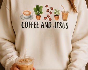 May include: A cream-coloured sweatshirt featuring the words "COFFEE AND JESUS" in black. The design includes a coffee cup, potted plants, coffee beans, and an iced coffee. Someone is holding an iced coffee.