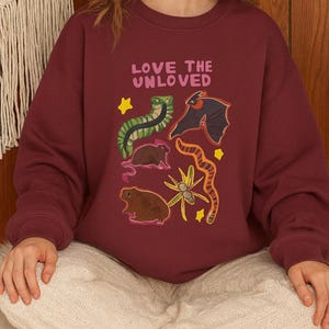May include: Burgundy sweatshirt with the text "LOVE THE UNLOVED" and illustrations of a snake, bat, and other creatures. The design includes yellow stars and a floral element. A cozy, casual top.