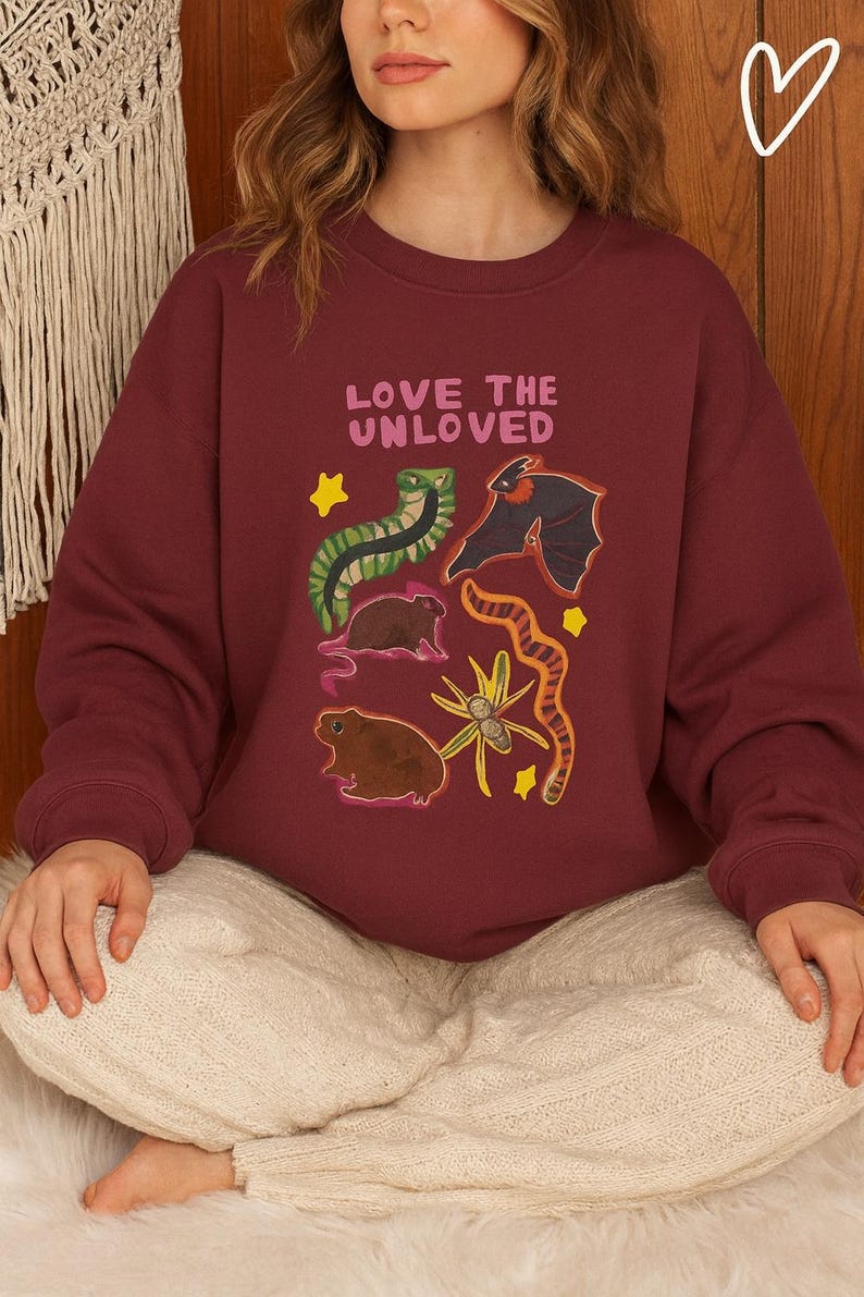 May include: Burgundy sweatshirt with the text "LOVE THE UNLOVED" and illustrations of a snake, bat, and other creatures. The design includes yellow stars and a floral element. A cozy, casual top.