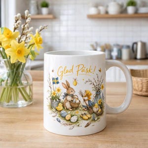 May include: White ceramic mug with a handle, featuring an Easter-themed illustration. The design includes a bunny, a girl, eggs, and the text "Glad Pask!" in gold lettering. The mug is decorated with spring flowers and a Swedish flag.