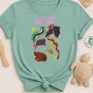 May include: A light teal t-shirt with the phrase "LOVE THE UNLOVED" in pink. The shirt features colorful illustrations of various creatures. A small turtle plush toy and a wooden brush are visible.