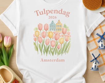 May include: White t-shirt with the text "Tulpendag 2026 Amsterdam" above a watercolor illustration of tulips and houses. The design includes pink and yellow tulips and green leaves. The shirt is styled with clogs and stroopwafels.