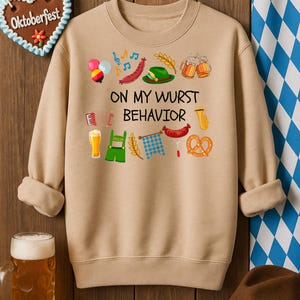 May include: Beige crew neck sweatshirt with the text "ON MY WURST BEHAVIOR" and Oktoberfest-themed graphics. Features beer steins, pretzels, sausages, and a green hat. A gingerbread heart with "Oktoberfest" is in the background.