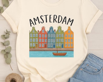 May include: Cream-colored t-shirt featuring a colorful illustration of Amsterdam canal houses and a small boat. The word "AMSTERDAM" is printed above the houses in black. The shirt is paired with blue jeans.