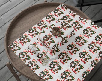 May include: A gift wrapped in white paper with a repeating pattern of the numbers "67" and the word "Seven" in red and green. A brown ribbon and bow adorn the gift. Scissors and tape are on the table.