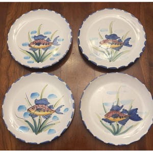 May include: A set of four round white ceramic plates featuring a hand-painted design of a fish in shades of orange, yellow, and blue, surrounded by green aquatic plants and blue water accents, with a scalloped blue rim around the edge.