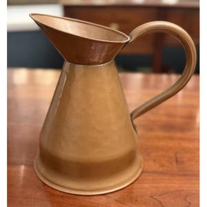 May include: A copper-colored metal pitcher with a wide, flared spout and a curved handle. The body of the pitcher features a distinct hammered texture, creating a series of subtle indentations across the surface, with a smooth, wide base.