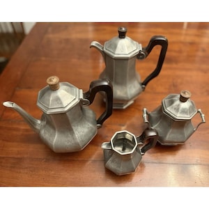 May include: A four-piece tea and coffee service set crafted from brushed pewter with a faceted, octagonal design. The set includes a tall coffee pot, a teapot, a lidded sugar bowl, and a creamer, all featuring dark brown handles and knobs.