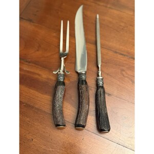 May include: A three-piece carving set consisting of a two-pronged meat fork, a sharp carving knife, and a sharpening steel. Each utensil features a textured, dark brown antler handle with a decorative silver-toned metal bolster at the base.