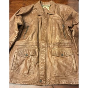 May include: A tan leather jacket featuring a collared neckline, a central button-down placket, two vertical zippered chest pockets, and two large lower patch pockets with buttoned flaps. The garment has a visible texture and a light-colored lining.
