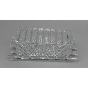 May include: A clear glass butter dish featuring a rectangular lid with vertical ribbed detailing and a base with a sunburst pattern radiating from the center. The glass has a faceted texture that catches light, creating a reflective appearance.