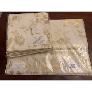 May include: A set of four cream-colored napkins and a set of four matching placemats, both featuring a metallic gold floral pattern. The items are packaged in clear plastic and include labels reading 18x18 NAPKINS and LA TABLE D'OR CHAMPAGNE.