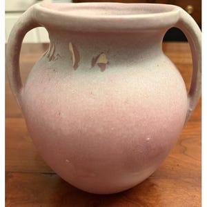 May include: A matte pink ceramic vase with a rounded body and two symmetrical handles extending from the rim to the shoulder. The surface features a soft gradient transition to a pale mint green hue near the top, with visible areas of paint wear.