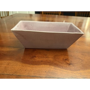 May include: A rectangular ceramic bowl with a light lavender glaze and a vertical ribbed texture on the exterior. The vessel features a wide, open top with angled sides that taper down to a smaller base, creating a clean, geometric silhouette.
