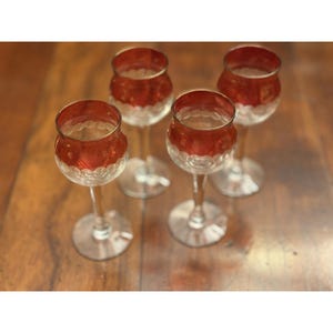 May include: A set of four stemmed glasses featuring a deep red upper bowl with a clear glass base. Each glass has a distinct scalloped or circular pattern etched around the lower section of the red bowl, supported by a slender clear stem and foot.