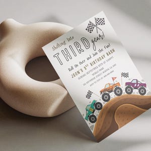 May include: A birthday party invitation with the text "Shifting Into Third Gear!" and "Jason's 3rd Birthday Bash." The invitation features illustrations of monster trucks racing on a dirt track, with checkered flags. A beige, donut-shaped sculpture is in the background.