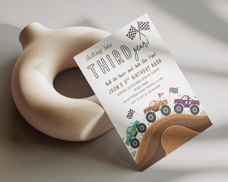 May include: A birthday party invitation with the text "Shifting Into Third Gear!" and "Jason's 3rd Birthday Bash." The invitation features illustrations of monster trucks racing on a dirt track, with checkered flags. A beige, donut-shaped sculpture is in the background.