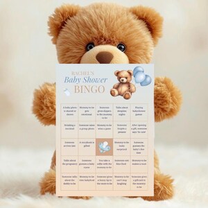Op de afbeelding: Een bruine teddybeer houdt een babyshower bingokaart vast. De kaart heeft een lichtblauw en wit ontwerp met de tekst "RACHEL'S Baby Shower BINGO" en een teddybeer-afbeelding. De bingokaart bevat verschillende babygerelateerde zinnen.