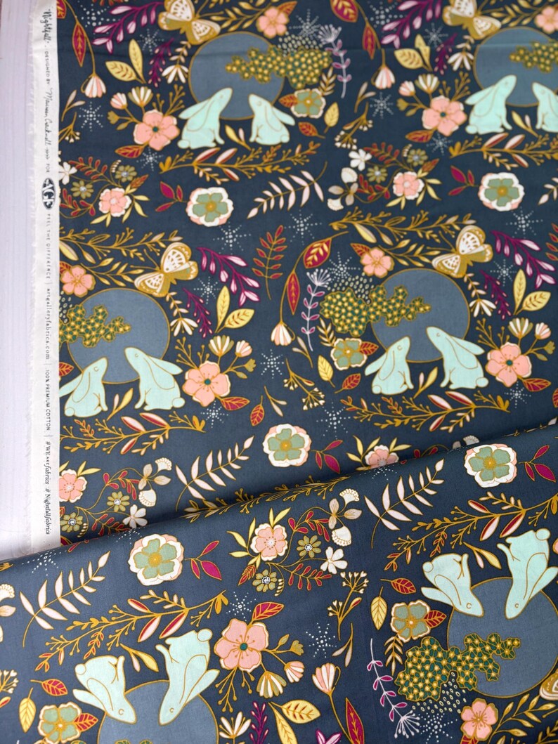 May include: Dark blue fabric with a repeating pattern of rabbits, butterflies, flowers, and foliage in shades of pink, green, gold, and white. The design includes a moon-like circle with two rabbits. The fabric is likely cotton.