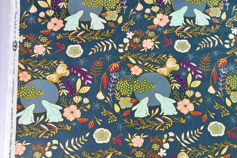 May include: Dark teal fabric with a repeating pattern of rabbits, flowers, butterflies, and foliage. The design includes light blue rabbits, pink and gold flowers, and gold butterflies. The fabric is 100% premium cotton.
