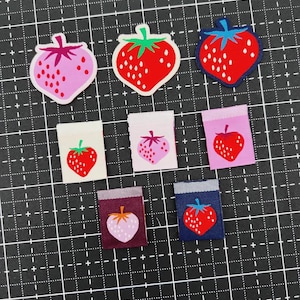 May include: A collection of seven fabric labels and patches featuring strawberry illustrations. Three are shaped like strawberries in pink, red, and dark blue, while four are rectangular with red or pink strawberries on white, pink, or dark backgrounds.