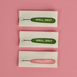 May include: Three rectangular fabric labels with a cream background. Each label features an embroidered green or pink oval shape containing the white text WELL, SHIT. A thin black line with a small red dot extends from the left side of the oval.