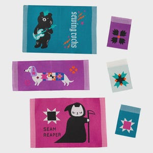 May include: A collection of six woven fabric labels featuring various designs: a black bear playing guitar with the text sewing rocks, a dachshund with a quilt pattern, a cat dressed as a grim reaper with the text SEAM REAPER, and three labels with geometric star motifs.