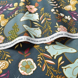 May include: Close-up of a fabric with a whimsical floral and bunny design. The fabric features light blue bunnies, pink and yellow flowers, and gold accents on a dark teal background. The text "100% Premium Cotton" is visible.