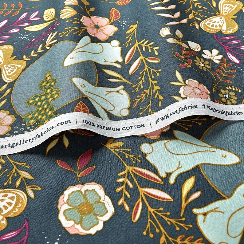 May include: Close-up of a fabric with a whimsical floral and bunny design. The fabric features light blue bunnies, pink and yellow flowers, and gold accents on a dark teal background. The text "100% Premium Cotton" is visible.