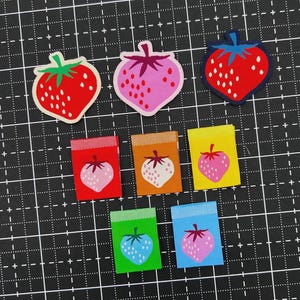 May include: A collection of eight fabric patches featuring strawberry designs. Three are shaped like strawberries in red, pink, and red with blue borders. Five are rectangular labels in red, orange, yellow, green, and blue, each with a strawberry icon.