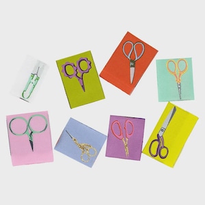 May include: Eight rectangular fabric labels featuring embroidered scissor designs in various colors including green, purple, orange, light blue, pink, and yellow. Each label displays a unique pair of scissors, showcasing different styles and handle shapes.