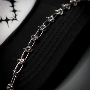 Könnte beinhalten: Nahaufnahme einer silberfarbenen Halskette mit Stacheldraht-Design. Die Kette liegt auf einem schwarzen Stoff, im Hintergrund ist eine schwarz-weiße Grafik zu sehen. Der Schmuck hat eine gotische oder Punk-Ästhetik.