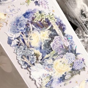 May include: A rectangular sticker sheet featuring a detailed watercolor illustration of a forest scene with a white winged deer, a full moon, blue hydrangea flowers, and white birds. The design includes holographic accents and various celestial elements.