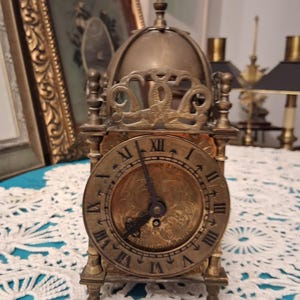 May include: Antique brass clock with a domed top and Roman numeral clock face. The clock has ornate detailing and stands on four small feet. The hands point to approximately 6:30. The clock is likely from the early 20th century.