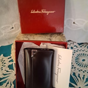 May include: A dark brown leather key case with a silver-tone metal F logo accent, presented in a red Salvatore Ferragamo branded box with white tissue paper and a matching informational card featuring the Salvatore Ferragamo brand name.
