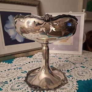 May include: A silver-colored, footed bowl with an ornate, scalloped rim and decorative cutouts. The bowl has a tall, slender stem and a wide, detailed base. The bowl is likely made of metal and reflects the surrounding environment.