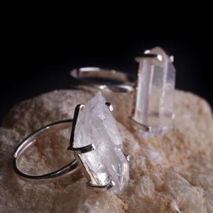 May include: Two silver rings with clear quartz crystal stones. The crystals are rough-cut and mounted on adjustable silver bands. The rings are displayed on a light-colored, textured surface against a dark background.