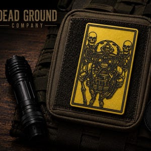May include: A rectangular yellow morale patch featuring a black graphic of a skeleton wearing tactical gear and night vision goggles, flanked by two smaller skulls. The patch is attached to a brown tactical pouch next to a black flashlight.