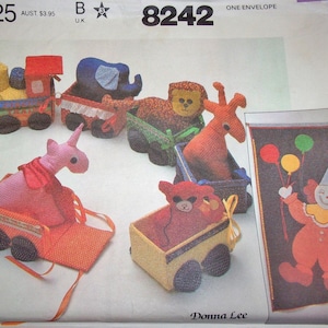 May include: A vintage McCall's sewing pattern featuring stuffed animal train cars and a clown banner. The train cars include a train engine, elephant, lion, giraffe, unicorn, and bear. The clown banner has balloons and the name "Jenny".