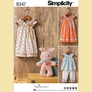 May include: Simplicity sewing pattern 8347. The image shows three children's outfits and a stuffed bunny. The outfits include a strawberry print dress, an orange dress with a white pattern, and a blue floral top with white pants. The bunny is pink with a heart.