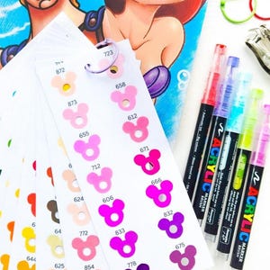 Puede incluir: Tarjetas de muestra de color con formas de cabeza de Mickey Mouse, con varios tonos de rosa, morado y marrón. Se ven rotuladores acrílicos en rojo, morado, azul, verde y amarillo. Ilustraciones de personajes de Disney en el fondo.