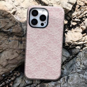 May include: A pink phone case with a white floral lace pattern. The case has a black camera lens frame and is resting on a rocky surface. The case is designed to fit a smartphone.
