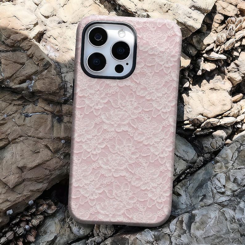 May include: A pink phone case with a white floral lace pattern. The case has a black camera lens frame and is resting on a rocky surface. The case is designed to fit a smartphone.