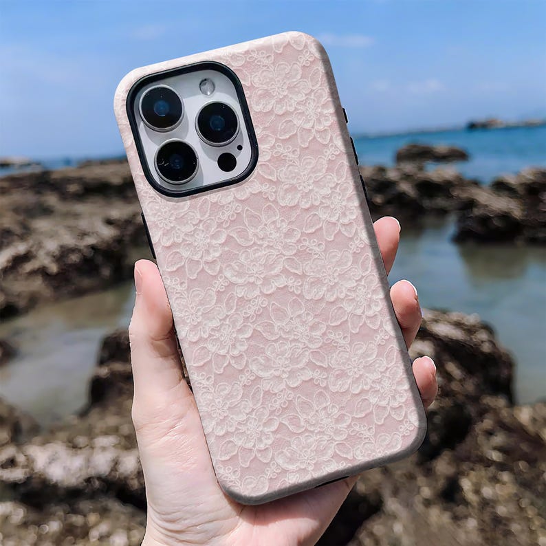 May include: A pink phone case with a white floral lace pattern. The case has a black border and a camera cutout. The background shows a beach with rocks and blue water.