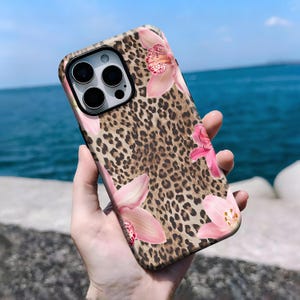 May include: A phone case featuring a leopard print design with pink orchid flower accents. The case has a black camera lens cutout and is held against a backdrop of the ocean and sky.