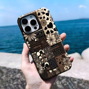 May include: A smartphone case featuring a patchwork design of black and beige squares. The pattern includes various floral motifs, polka dots, grid lines, and heart shapes. The case has a cutout for a triple-lens camera system on the back.